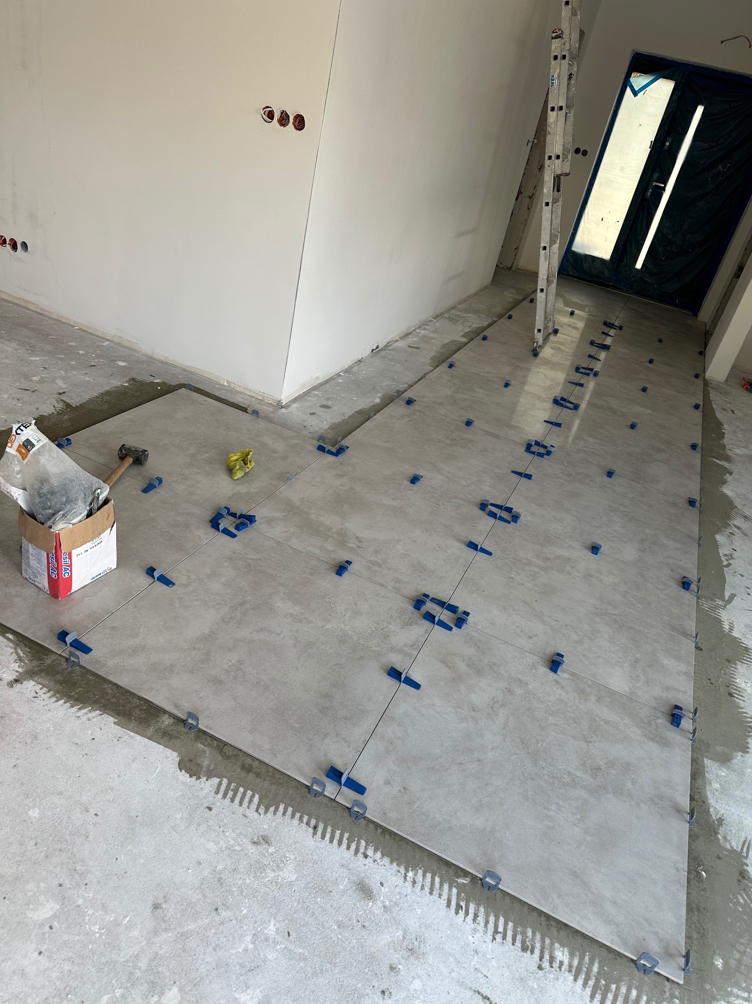 Układanie szarych płytek podłogowych z użyciem niebieskich klipsów poziomujących, widoczna drabina oparta o ścianę i drzwi zabezpieczone folią, karton z zaprawą klejową i gumowy młotek.
