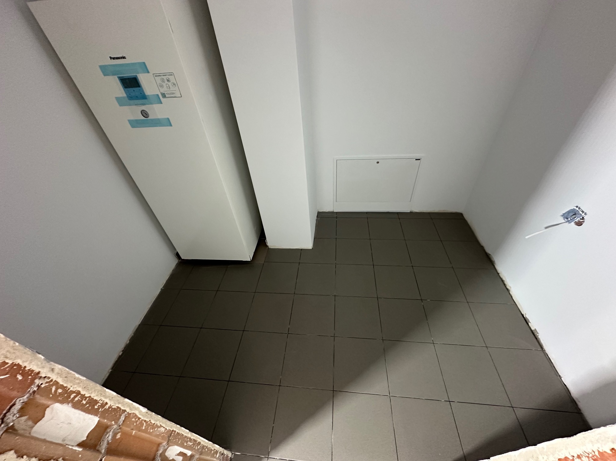 Wnętrze pomieszczenia z szarą, kwadratową podłogą z płytek, białą ścianą z widoczną instalacją elektryczną i białą obudową pompy ciepła Panasonic, widoczne fragmenty surowej cegły u dołu kadru.