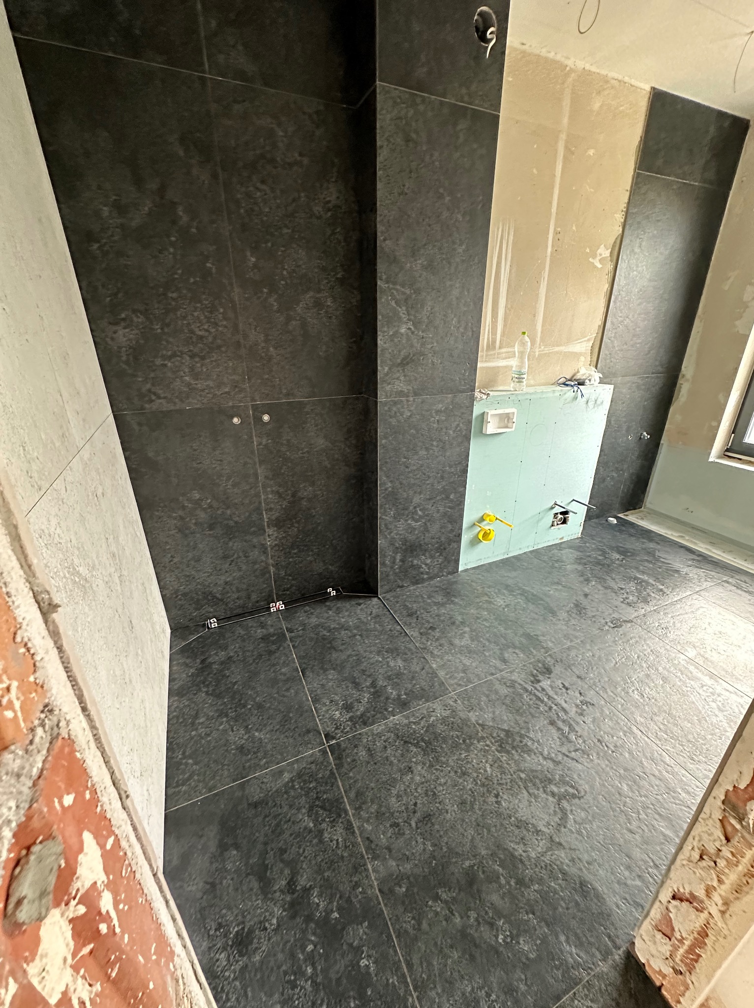 Częściowo wykończona łazienka z ciemnymi płytkami na ścianach i podłodze, widoczne elementy instalacji wodnej i fragment ściany z płyt gipsowo-kartonowych przygotowany pod montaż stelaża...