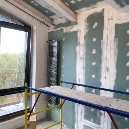 Aleksandra Szymborska CREO COMPANY - Wnętrze w trakcie szpachlowania ścian i sufitu płytami gipsowo-kartonowymi, widoczne łączenia płyt wypełnione masą szpachlową, rusztowanie malarskie i okno z widokiem na drzewa.