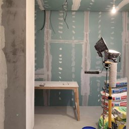 Aleksandra Szymborska CREO COMPANY - Ściana z płyt gipsowo-kartonowych w trakcie przygotowania do malowania, widoczne szpachlowanie łączeń, oświetlenie robocze, stół roboczy i worki z zaprawą gipsową