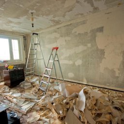 Aleksandra Szymborska CREO COMPANY - Pokój w trakcie remontu: zerwana tapeta, ściany przygotowane do szpachlowania, dwie drabiny, laptop i resztki tapety na podłodze, widoczny stary kredens pod oknem.