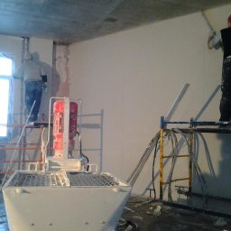Aleksandra Szymborska CREO COMPANY - Trzech mężczyzn na rusztowaniach aplikuje gładź na ścianę w surowym pomieszczeniu z betonowym sufitem, widoczna maszyna do gładzi z czerwonym silnikiem.