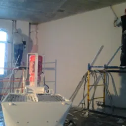 Trzech mężczyzn na rusztowaniach aplikuje gładź na ścianę w surowym pomieszczeniu z betonowym sufitem, widoczna maszyna do gładzi z czerwonym silnikiem.