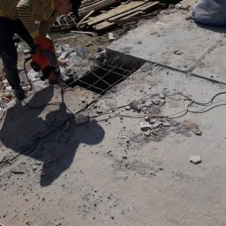 Aleksandra Szymborska CREO COMPANY - Prace remontowe: robotnik używa młota udarowego do wykuwania otworu w betonowej podłodze, widoczna siatka zbrojeniowa, gruz i szlifierka kątowa obok.