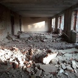 Aleksandra Szymborska CREO COMPANY - Wnętrze w trakcie rozbiórki: gruz, cegły i fragmenty betonu na podłodze, odsłonięte ściany z cegły i betonowe filary, wpadające światło przez okna.