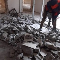 Aleksandra Szymborska CREO COMPANY - Demontaż posadzki w remontowanym pomieszczeniu; gruz i fragmenty płytek na podłodze, pracownik w kamizelce ochronnej używa młota udarowego.