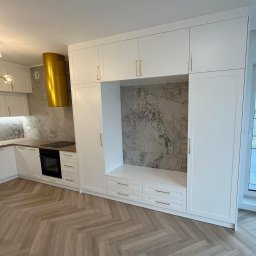 Aleksandra Szymborska CREO COMPANY - Nowoczesna kuchnia z białymi szafkami pod sufit, złotymi uchwytami, marmurowym backsplashiem i złotym okapem, drewniana podłoga ułożona w jodełkę.