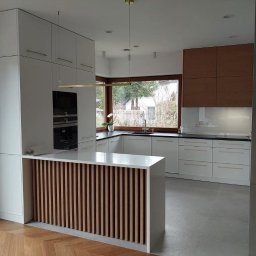 Aleksandra Szymborska CREO COMPANY - Nowoczesna kuchnia z białymi szafkami, drewnianymi akcentami i wyspą z dekoracyjnymi listwami, oświetlona wiszącą lampą i naturalnym światłem z okna.