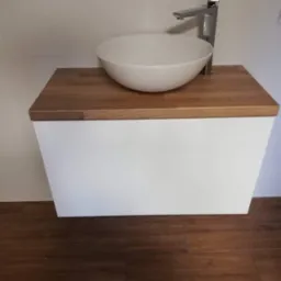 Nowoczesna umywalka nablatowa w okrągłym kształcie, osadzona na drewnianym blacie szafki z białym frontem, minimalistyczna bateria chromowana, płytki podłogowe imitujące drewno.