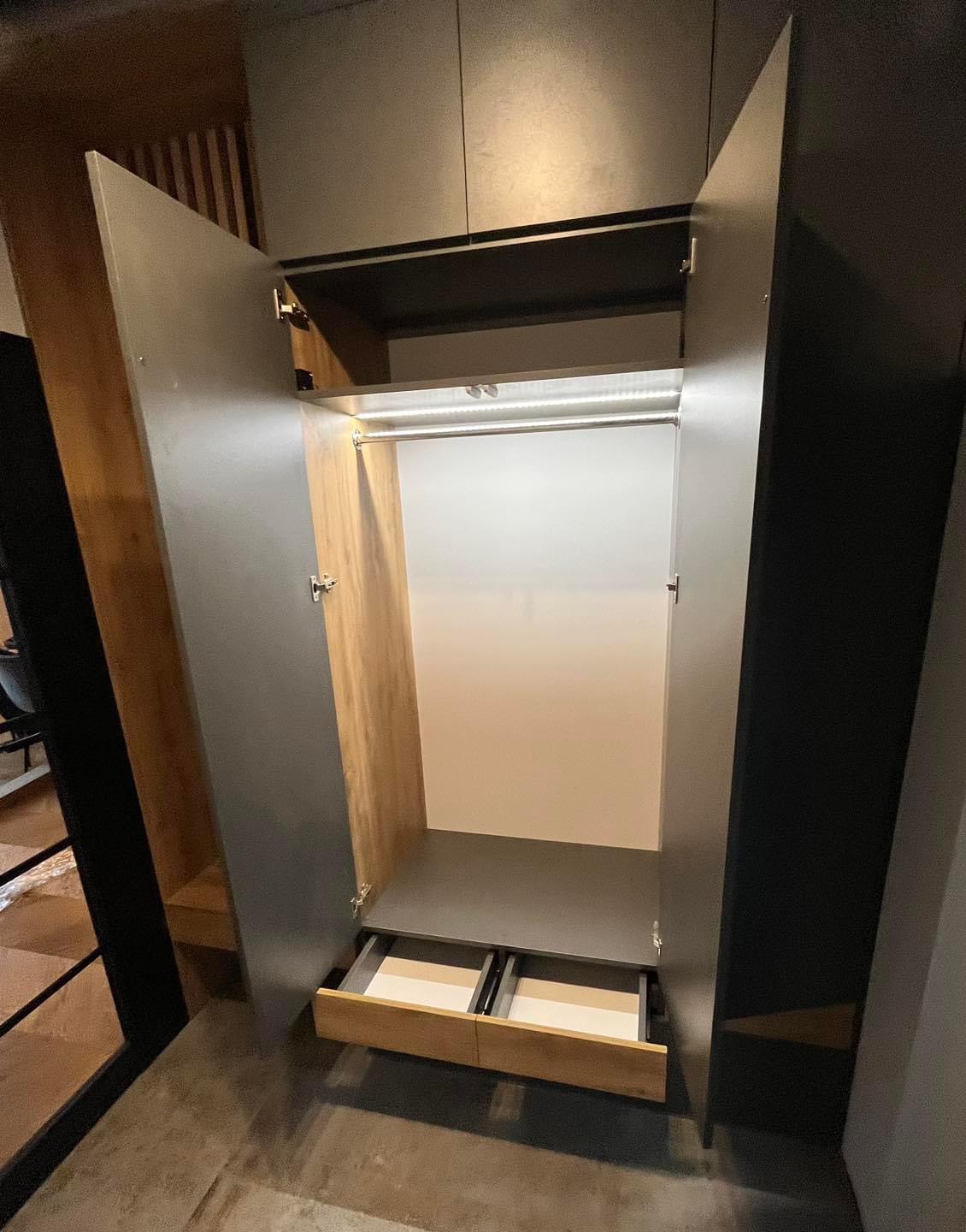 Otwarta szafa wnękowa z szarymi drzwiami, drewnianym wnętrzem i dwoma wysuwanymi szufladami na dole, oświetlona listwą LED.