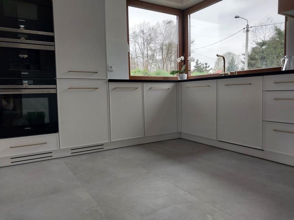 Nowoczesna, jasna kuchnia z białymi szafkami z minimalistycznymi, złotymi uchwytami, zabudowanym sprzętem AGD marki Siemens i dużym oknem z widokiem na drzewa.