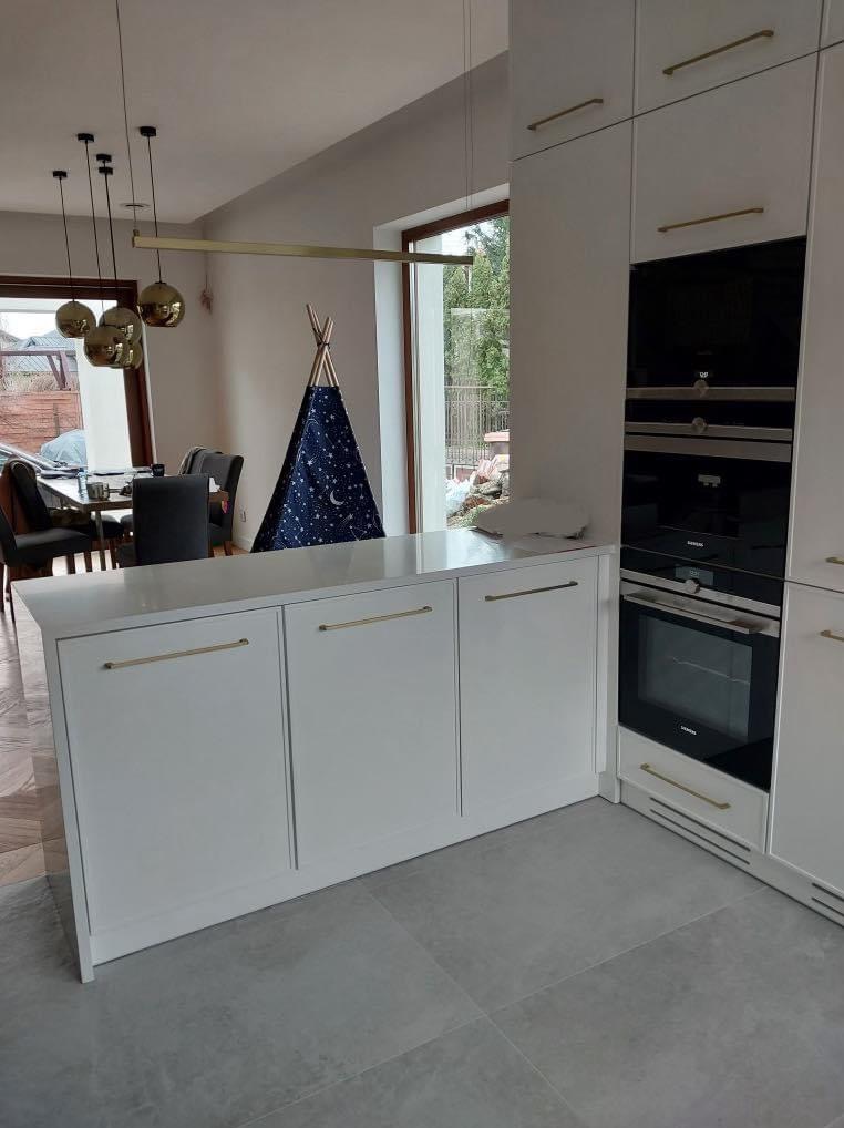 Nowoczesna, biała zabudowa kuchenna z złotymi uchwytami, widok na jadalnię z dekoracyjnym tipi w tle.