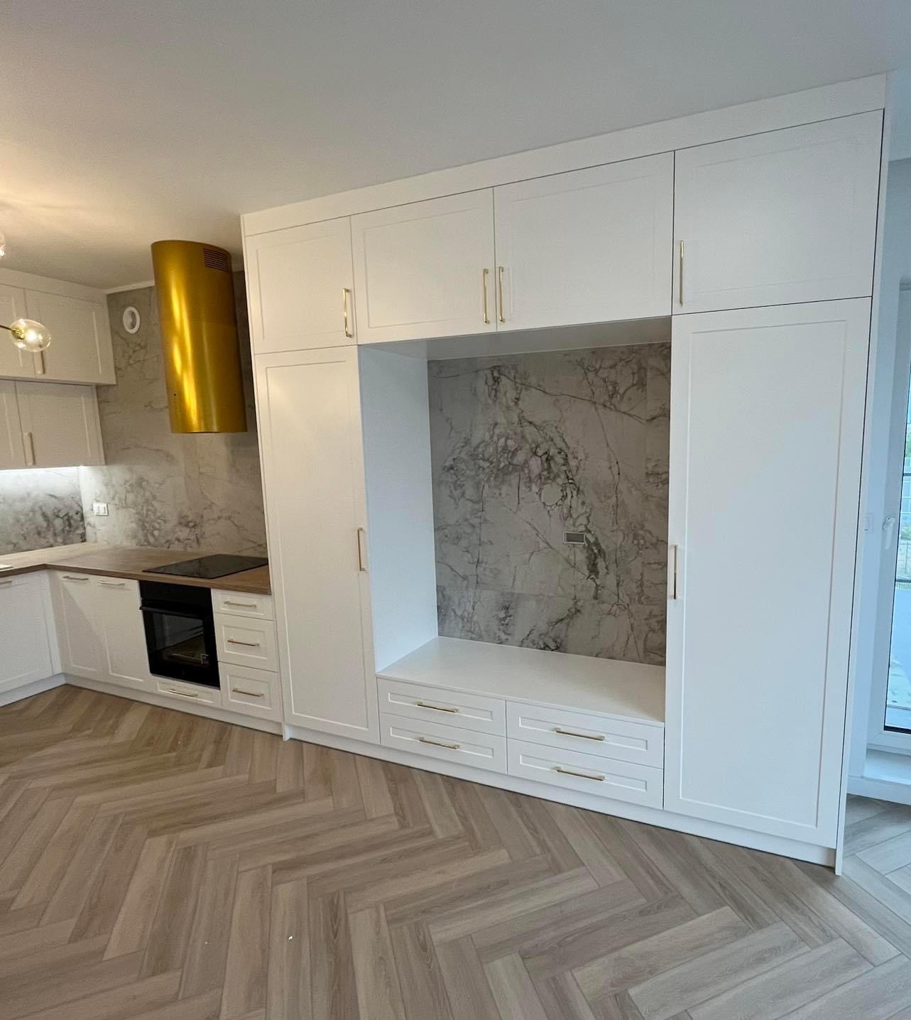 Nowoczesna kuchnia z białymi szafkami pod sufit, złotymi uchwytami, marmurowym backsplashiem i złotym okapem, drewniana podłoga ułożona w jodełkę.