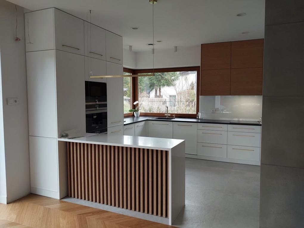 Nowoczesna kuchnia z białymi szafkami, drewnianymi akcentami i wyspą z dekoracyjnymi listwami, oświetlona wiszącą lampą i naturalnym światłem z okna.