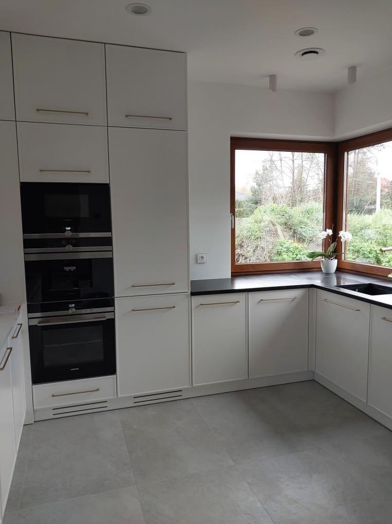 Jasna kuchnia z zabudową meblową do sufitu, trzema piekarnikami i narożnym oknem z widokiem na zieleń.