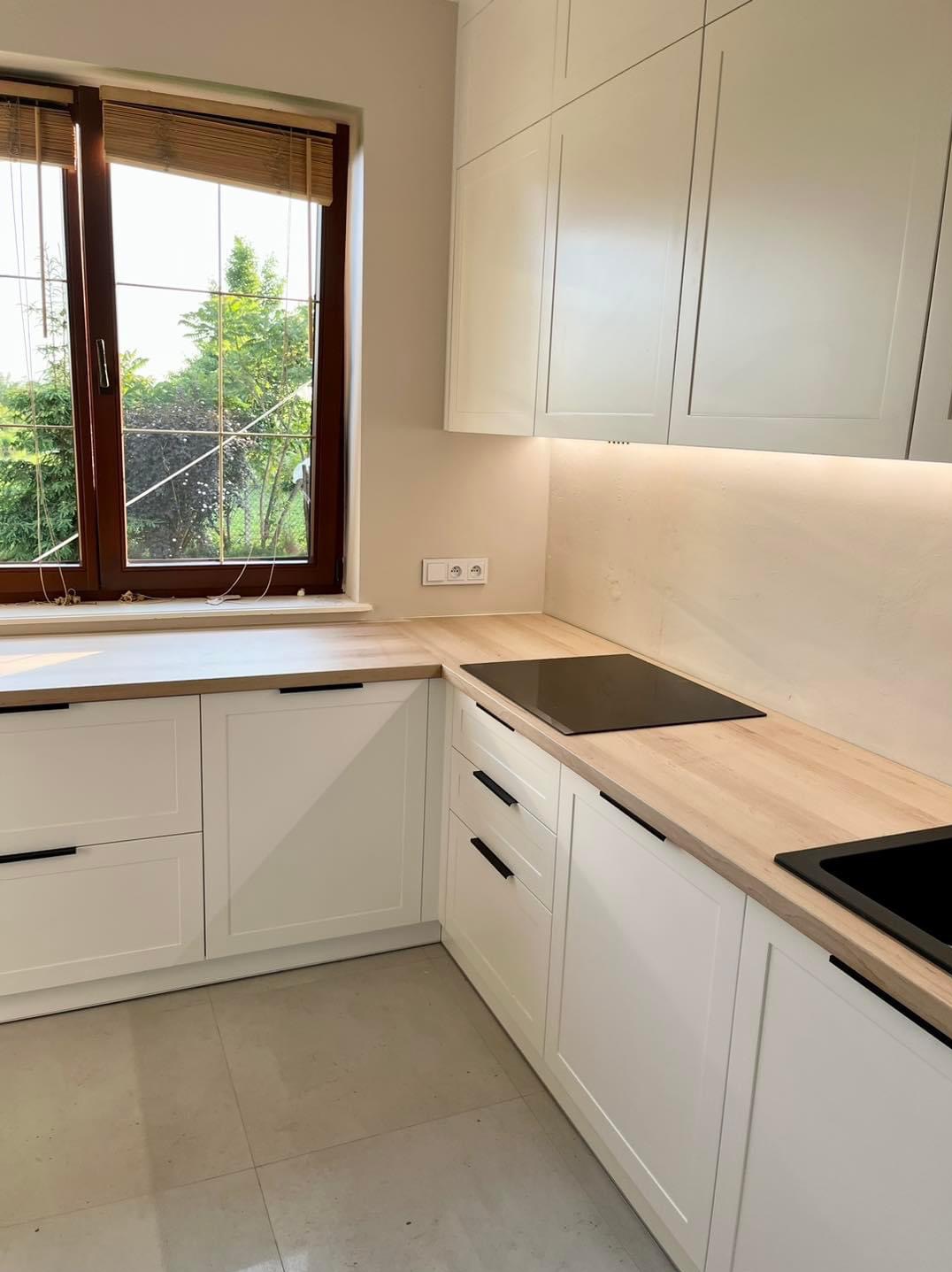 Nowoczesna kuchnia z białymi szafkami, drewnianym blatem i czarnymi uchwytami, widok na okno z roletami bambusowymi i zielenią za oknem.