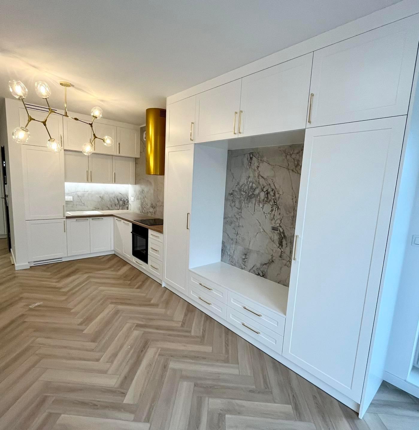 Nowoczesna, jasna kuchnia z białymi szafkami, marmurowym backsplashiem i złotym okapem, parkiet w jodełkę na podłodze, oświetlona designerską lampą sufitową.