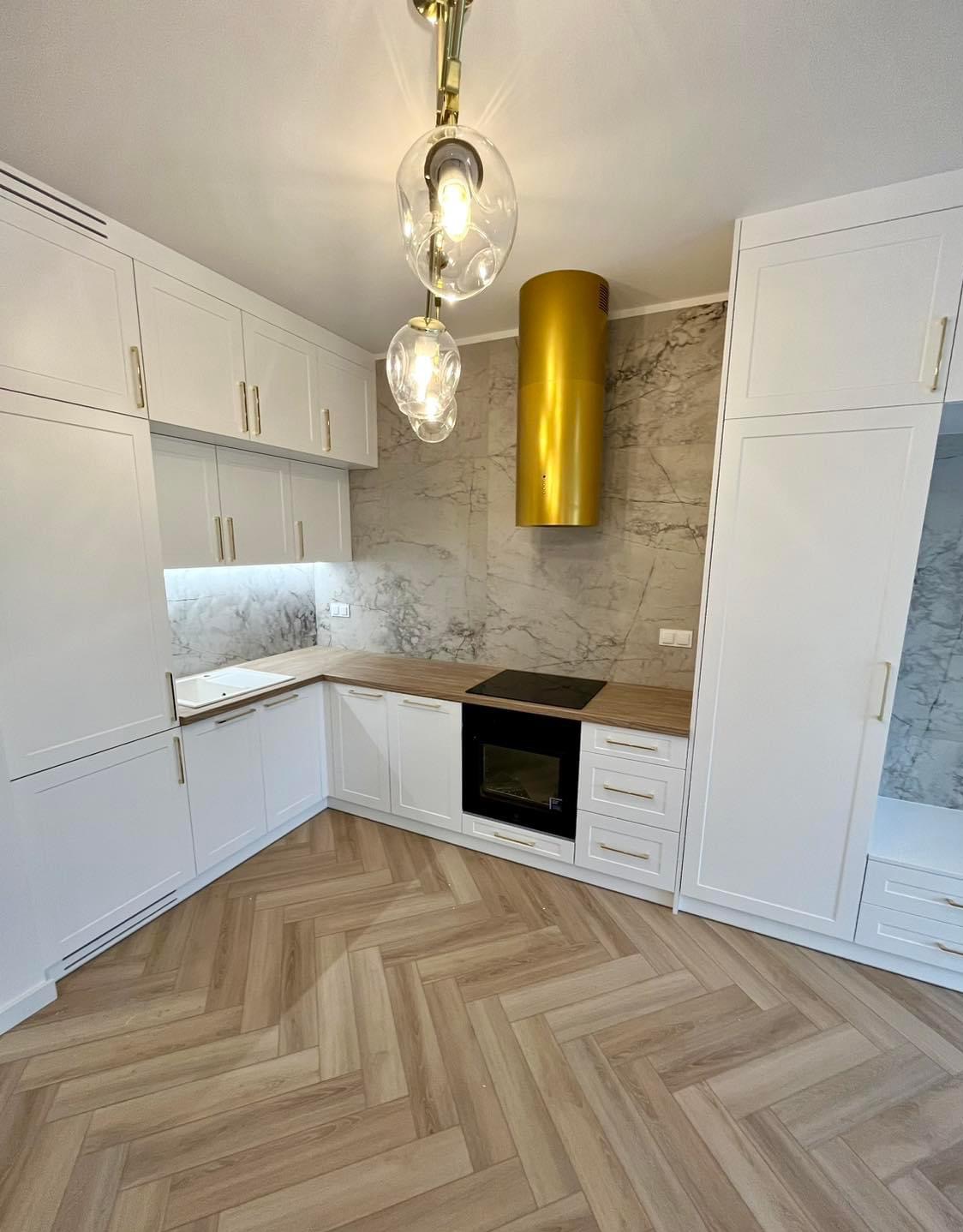 Nowoczesna kuchnia z białymi szafkami, drewnianym blatem i złotym okapem cylindrycznym, podłoga ułożona w jodełkę, oświetlenie punktowe pod szafkami i designerskie lampy wiszące.