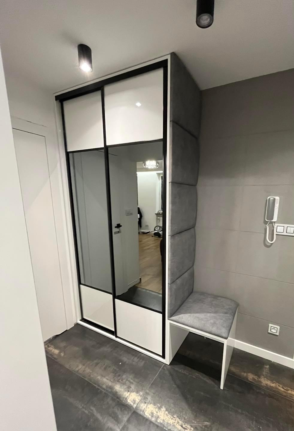 Nowoczesna szafa wnękowa z przesuwnymi drzwiami lustrzanymi, połączona z tapicerowanym siedziskiem i panelem ściennym w odcieniach szarości, tworząca funkcjonalną strefę wejściową w mieszkaniu.