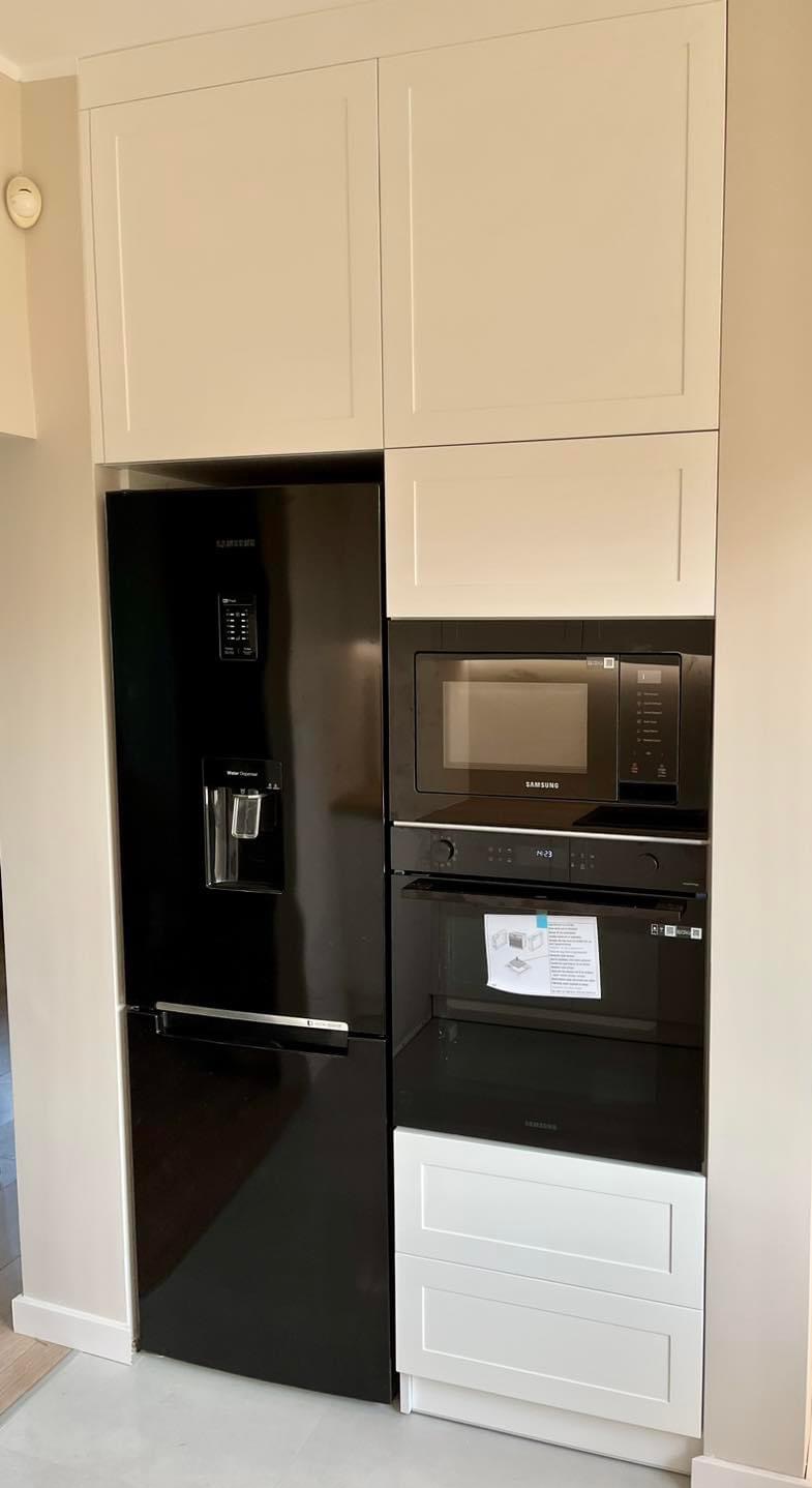 Nowoczesna zabudowa kuchenna z czarną lodówką Samsung z kostkarką, piekarnikiem i mikrofalówką w czarnych obudowach, uzupełniona białymi szafkami górnymi i dolnymi z frontami na wysoki połysk.