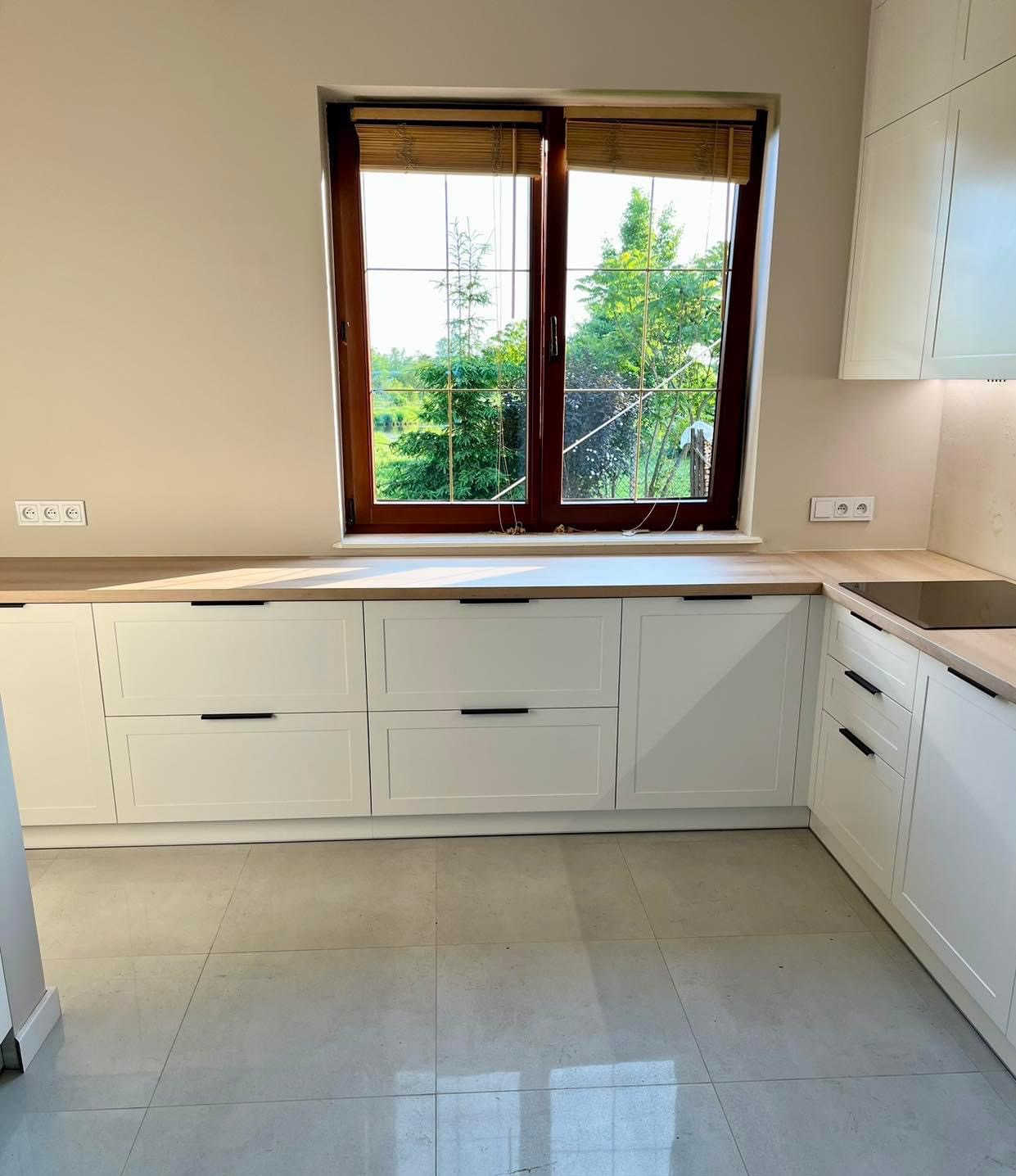 Jasna kuchnia z białymi szafkami z czarnymi uchwytami i drewnianym blatem, widok na zewnątrz przez okno z drewnianą ramą i bambusowymi roletami, płytki podłogowe w odcieniach szarości.