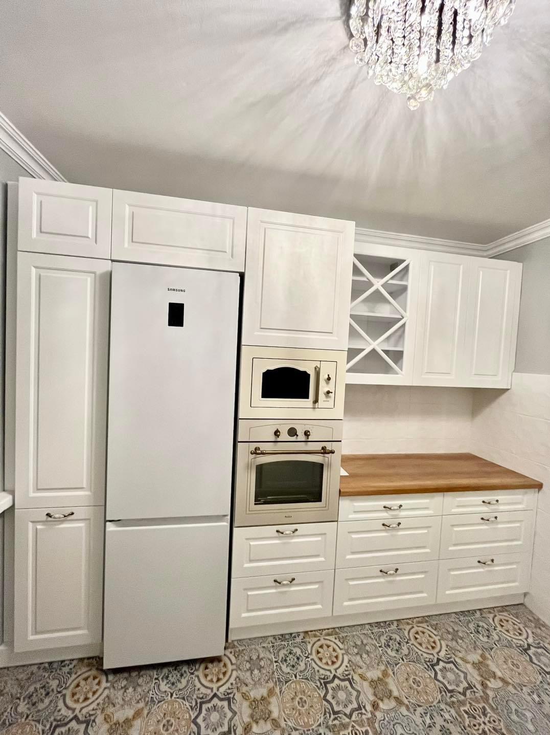 Biała kuchnia z zabudową meblową, w tym lodówka, piekarnik, mikrofalówka i szafki, uzupełniona drewnianym blatem i wzorzystą podłogą, oświetlona kryształowym żyrandolem.