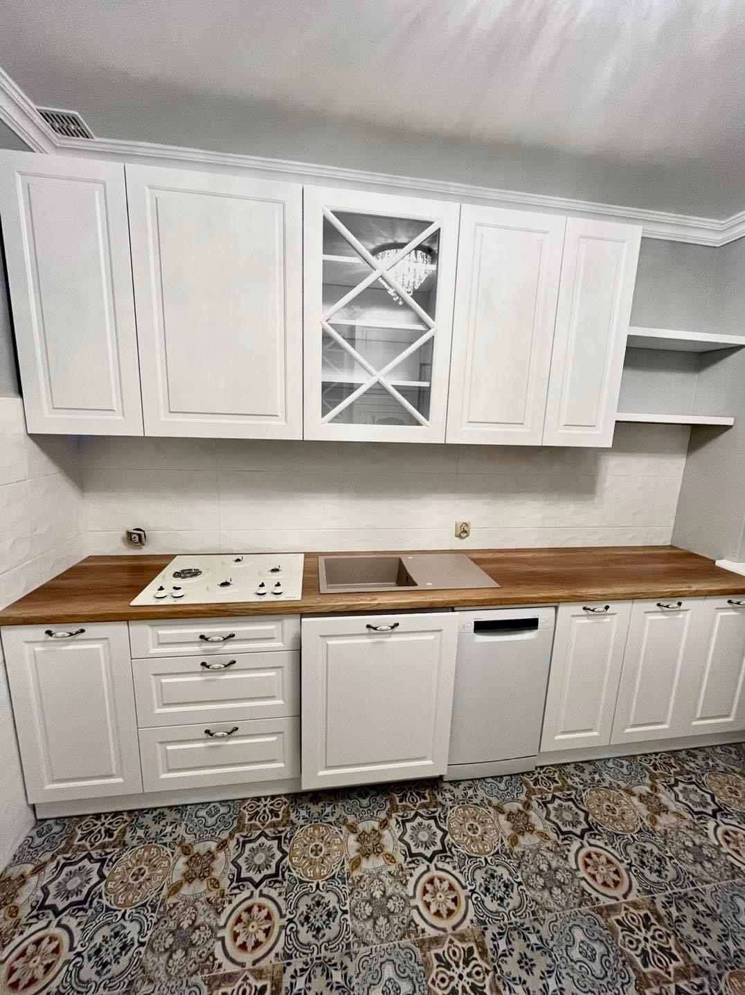 Biała zabudowa kuchenna z drewnianym blatem i wzorzystą podłogą. W górnych szafkach witryna z krzyżowymi szprosami, za którą widać fragment żyrandola.