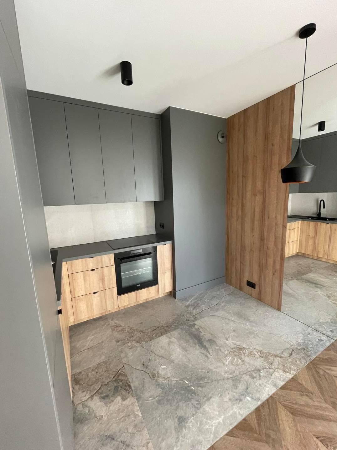 Nowoczesna kuchnia z szarymi szafkami wiszącymi i dolnymi szafkami z drewnianym fornirem, marmurowa podłoga i drewniana ścianka działowa, oświetlenie punktowe i designerska lampa wisząca.