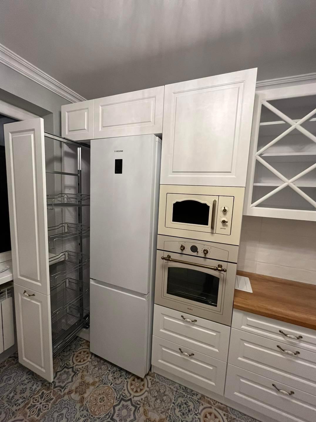 Biała kuchnia z zabudową, w której znajduje się lodówka Samsung, piekarnik Amica w odcieniach beżu i wysuwana szafka cargo z metalowymi koszami, na podłodze wzorzyste płytki.