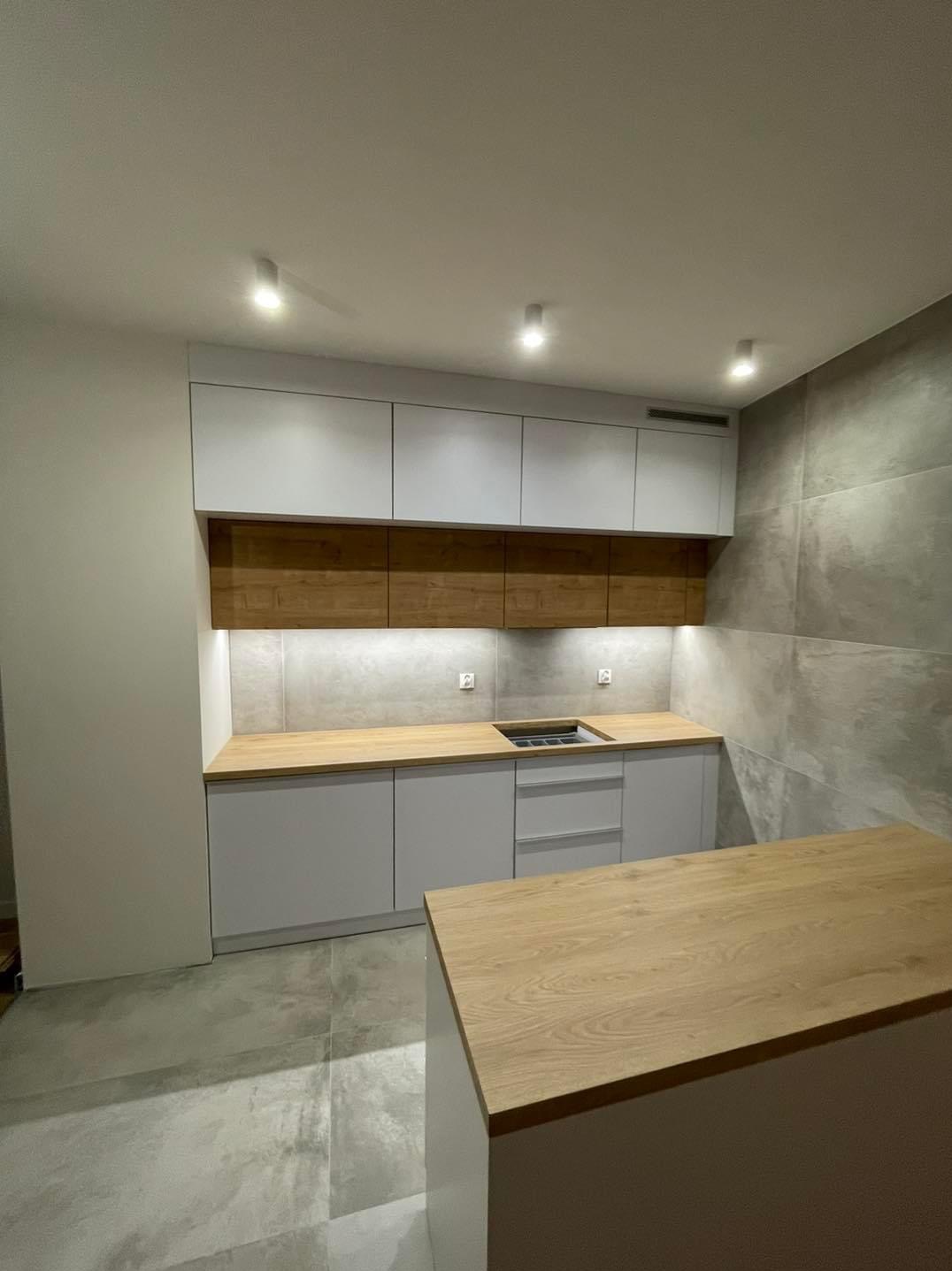 Nowoczesna kuchnia z szarymi szafkami dolnymi i białymi górnymi, drewnianym blatem i wyspą, oświetlona punktowo, z szarą płytką na ścianie.