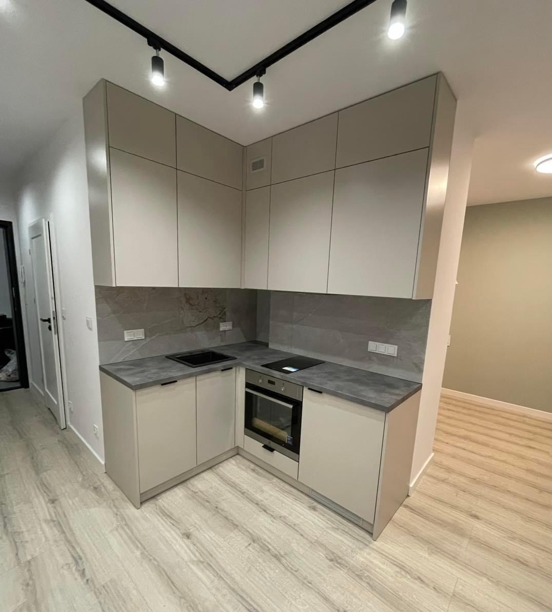 Nowoczesna, narożna kuchnia w odcieniach beżu i szarości w Żyrardowie. Górne szafki sięgają sufitu, a podświetlenie szynowe podkreśla minimalistyczny design. Blat z szarego kamienia kontrastuje...