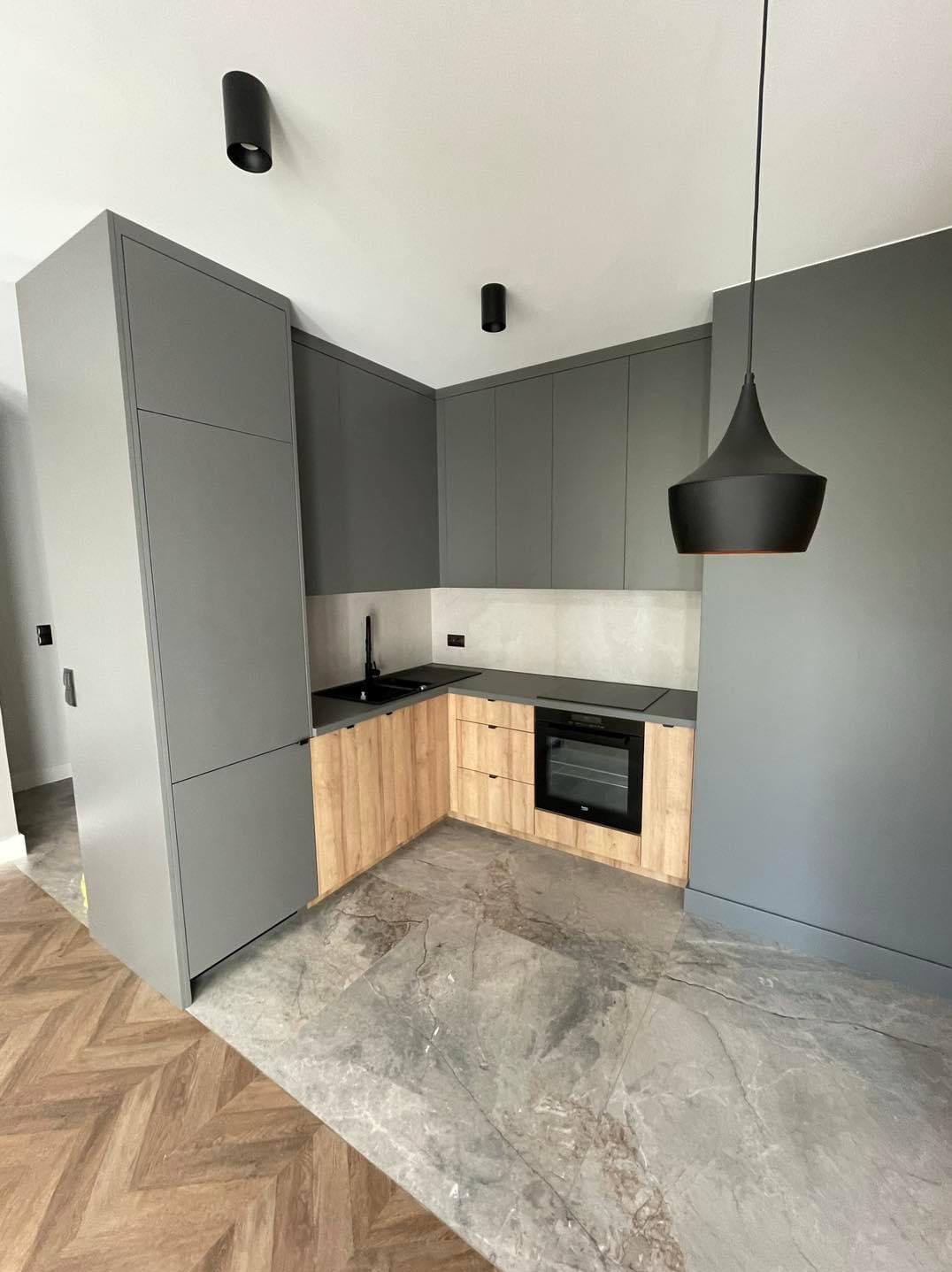 Nowoczesna kuchnia z szarymi szafkami i drewnianymi frontami, czarnym zlewem i płytą indukcyjną, podłoga z szarego marmuru i parkiet w jodełkę w przyległym pomieszczeniu, oświetlenie punktowe...