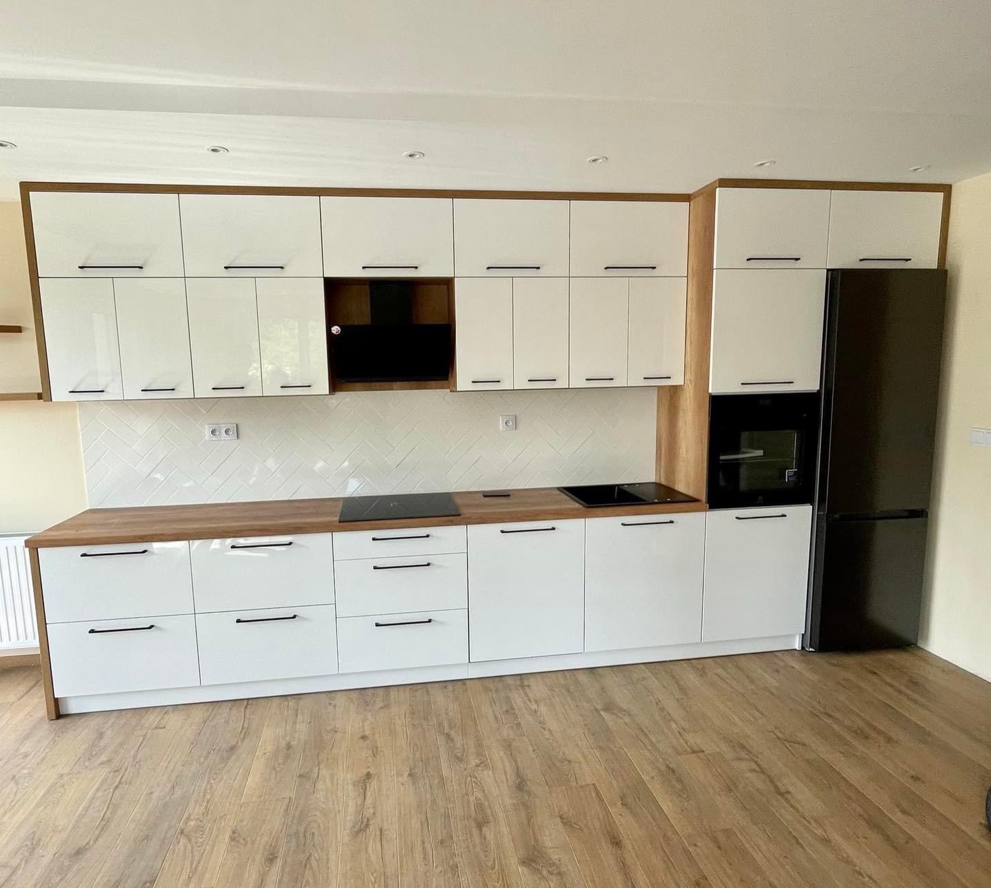 Nowoczesna, biała kuchnia z drewnianym blatem i czarnymi uchwytami, wykończona płytkami ułożonymi w jodełkę na ścianie i drewnianą podłogą.