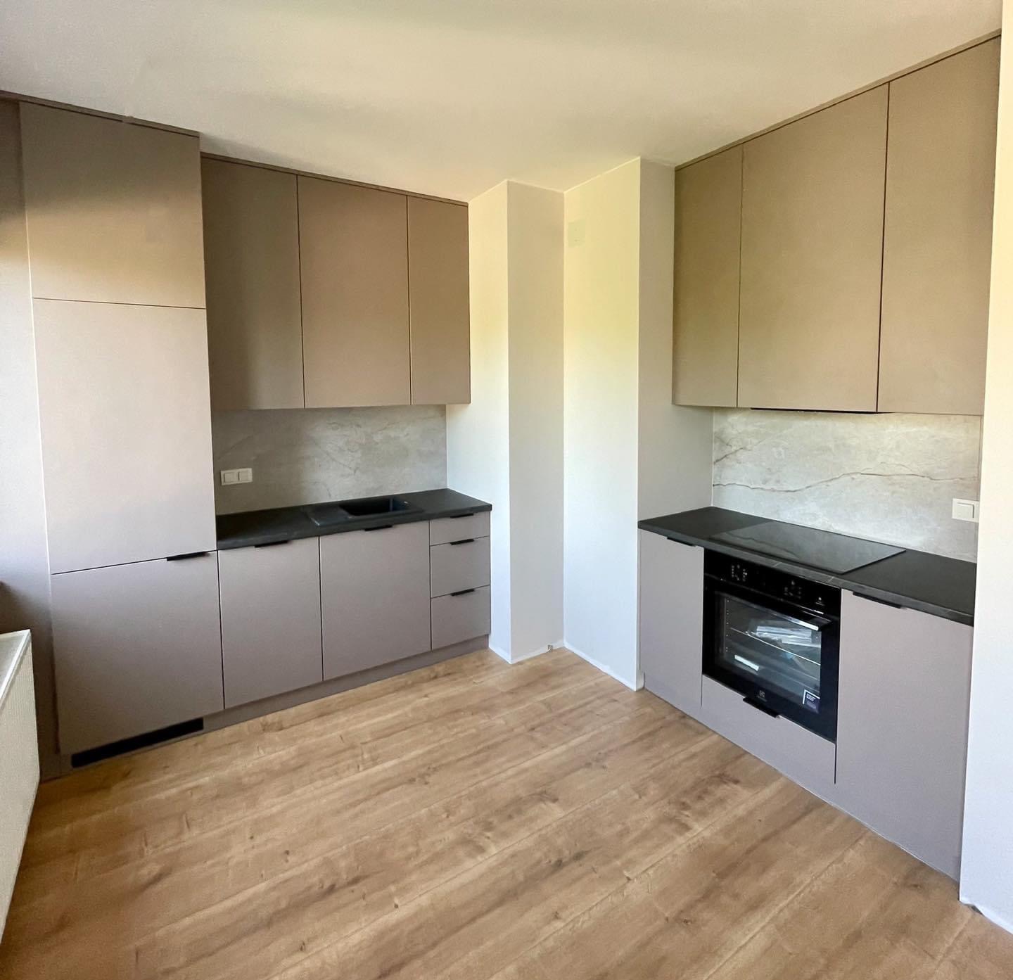 Nowoczesna kuchnia w odcieniach szarości i beżu z drewnopodobną podłogą, minimalistyczne szafki bez uchwytów, czarny blat i płyta indukcyjna, marmurowy backsplash.