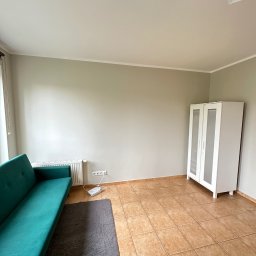 MADMAL - Jasnoszary pokój po remoncie z turkusową sofą, białym regałem z przeszkleniami i brązowymi płytkami podłogowymi, widoczny router WiFi i szary dywanik.