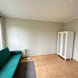 Jasnoszary pokój po remoncie z turkusową sofą, białym regałem z przeszkleniami i brązowymi płytkami podłogowymi, widoczny router WiFi i szary dywanik.