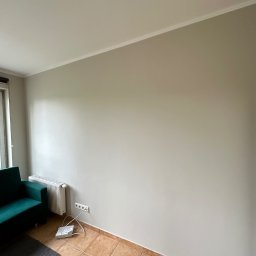 MADMAL - Jasnoszara ściana po remoncie, widoczne gniazdka elektryczne, grzejnik, fragment zielonego fotela i szary dywanik na brązowych płytkach podłogowych.