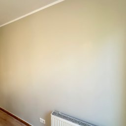 MADMAL - Ściana pomalowana na jasny, neutralny kolor z widocznym grzejnikiem, gniazdkiem elektrycznym i fragmentem podłogi z paneli. Widoczna listwa przypodłogowa i biała listwa sufitowa.