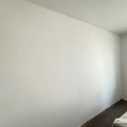 Świeżo pomalowana na biało ściana w pokoju z brązową wykładziną, widoczne zabezpieczenia podłogi przy ścianie.