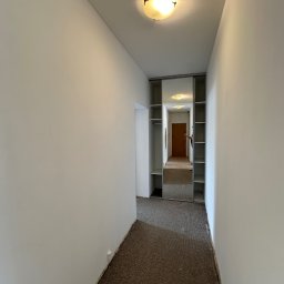 MADMAL - Korytarz w mieszkaniu po odświeżeniu ścian, widoczne ślady po niedokładnym zabezpieczeniu podłogi dywanowej przy listwach przypodłogowych i gładkie, białe powierzchnie ścian.