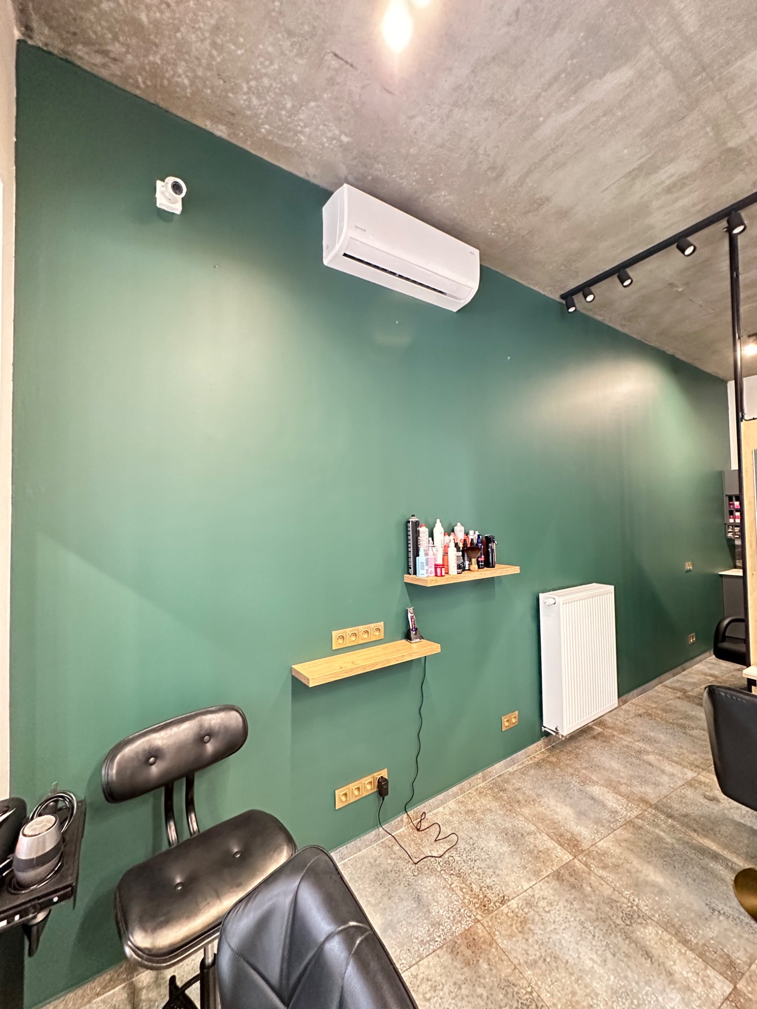 Ściana w odcieniach zieleni w salonie fryzjerskim, z drewnianymi półkami, grzejnikiem, klimatyzatorem i oświetleniem punktowym na suficie.