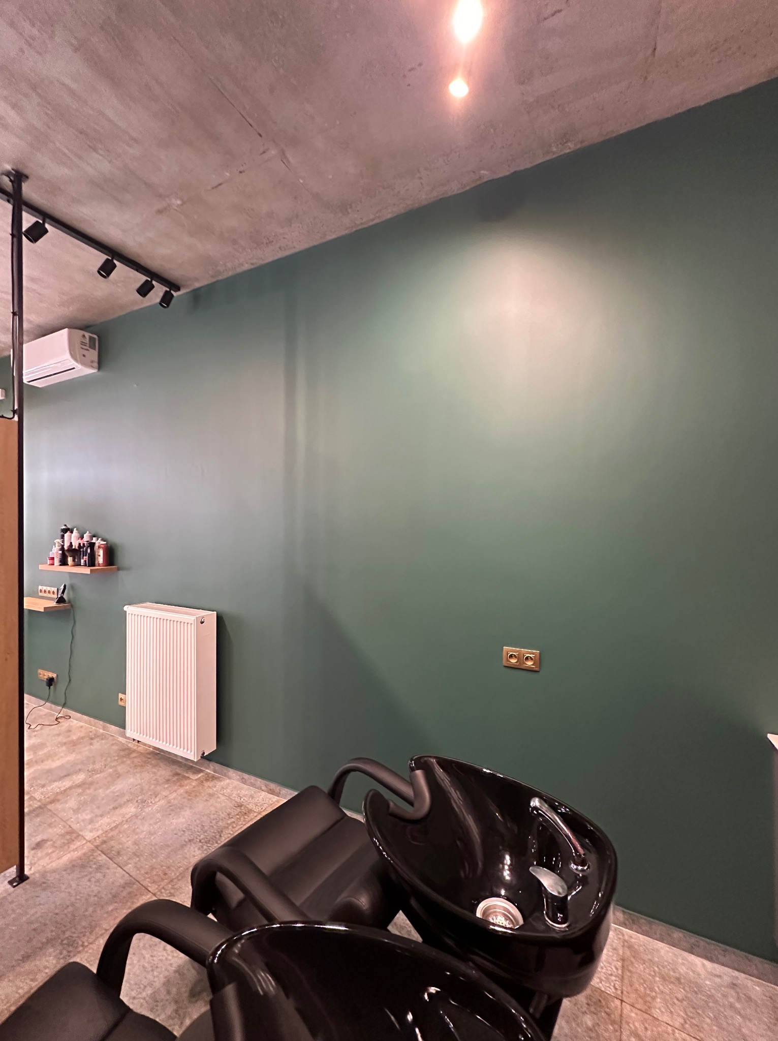 Nowoczesne wnętrze salonu fryzjerskiego z ciemnozieloną ścianą, betonowym sufitem z reflektorami, czarnym fotelem z myjnią oraz białym grzejnikiem.