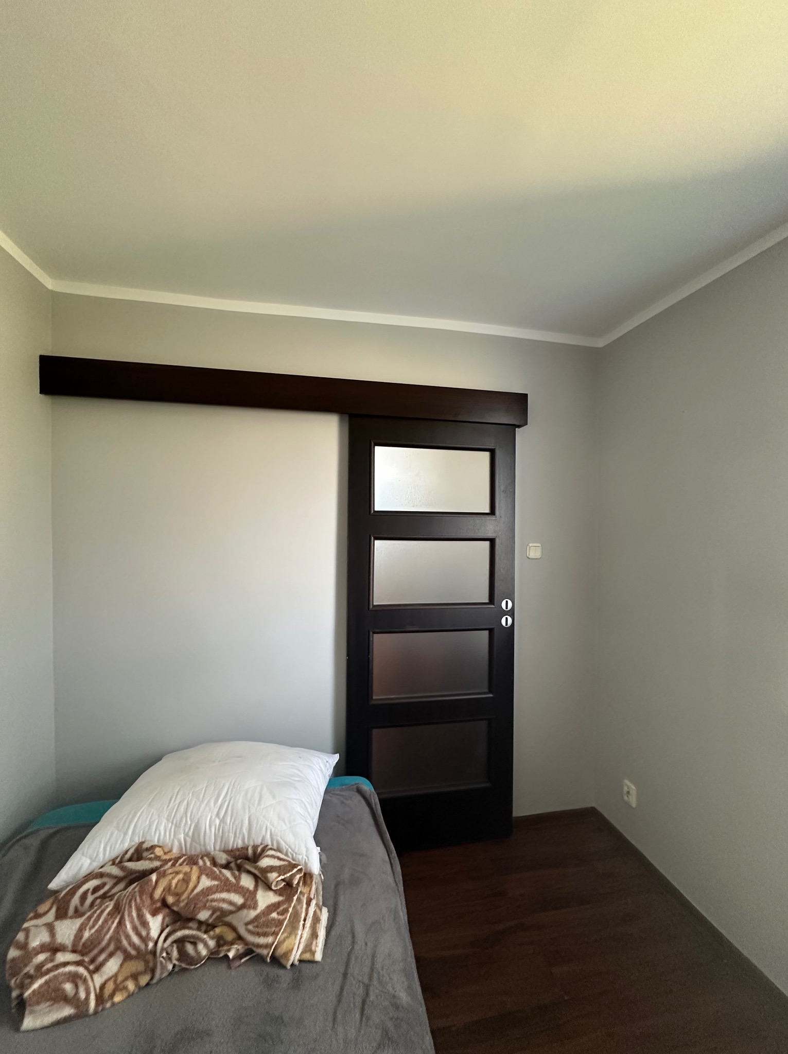 Pokój z szarą ścianą, ciemną przesuwną szklaną stolarką drzwiową oraz podłogą z ciemnego drewna, widoczny fragment łóżka z pościelą.