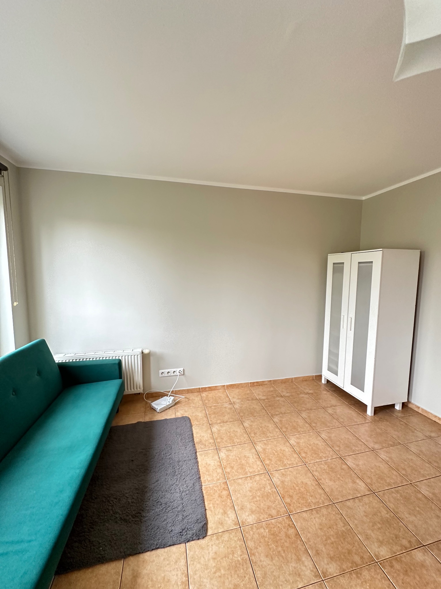 Jasnoszary pokój po remoncie z turkusową sofą, białym regałem z przeszkleniami i brązowymi płytkami podłogowymi, widoczny router WiFi i szary dywanik.