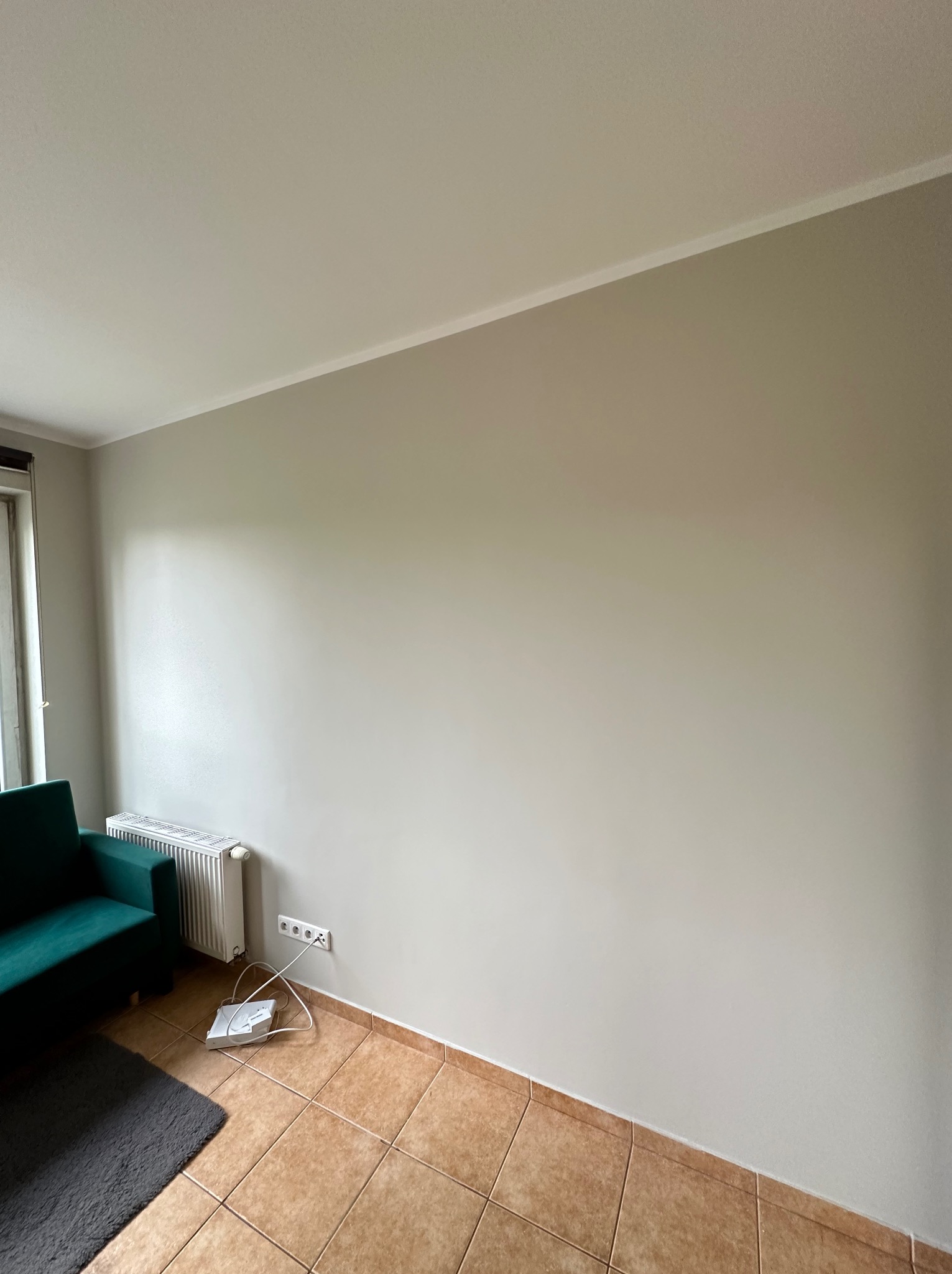 Jasnoszara ściana po remoncie, widoczne gniazdka elektryczne, grzejnik, fragment zielonego fotela i szary dywanik na brązowych płytkach podłogowych.