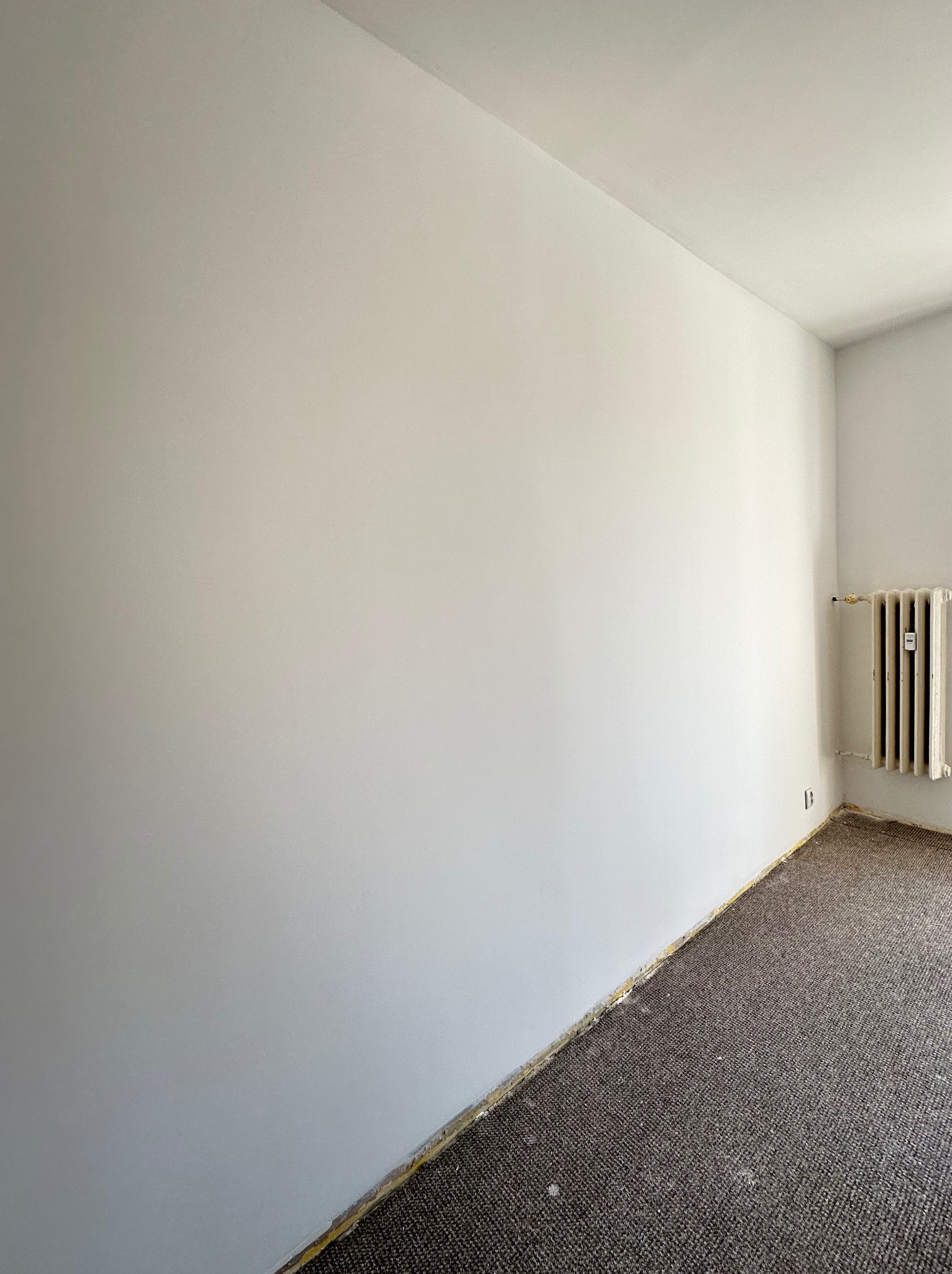 Jasnoszara, świeżo pomalowana ściana w pokoju z brązową wykładziną, widoczny stary grzejnik i listwy przypodłogowe zabezpieczone taśmą malarską.