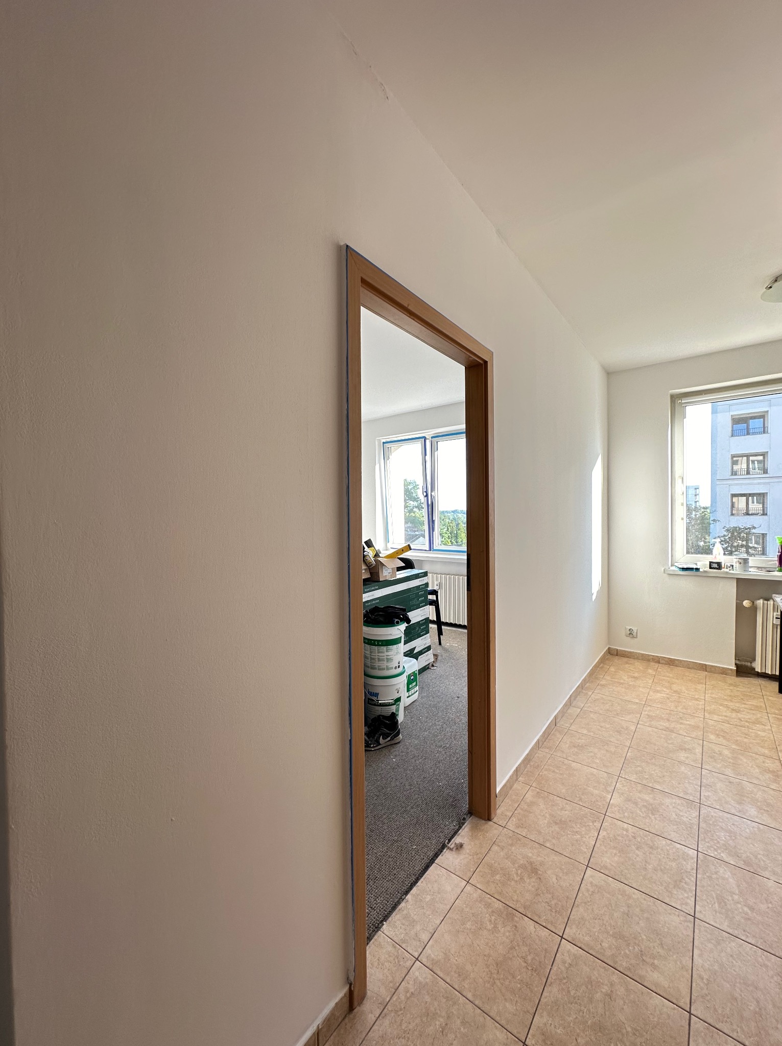 Świeżo pomalowane wnętrze mieszkania z widocznymi wiadrami z farbą w sąsiednim pokoju, jasne ściany i brązowe płytki podłogowe.