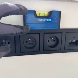 Sprawdzanie poziomu czarnej listwy z gniazdami elektrycznymi i portem USB za pomocą poziomicy Dexter przez osobę w rękawicy roboczej na tle białej ściany.