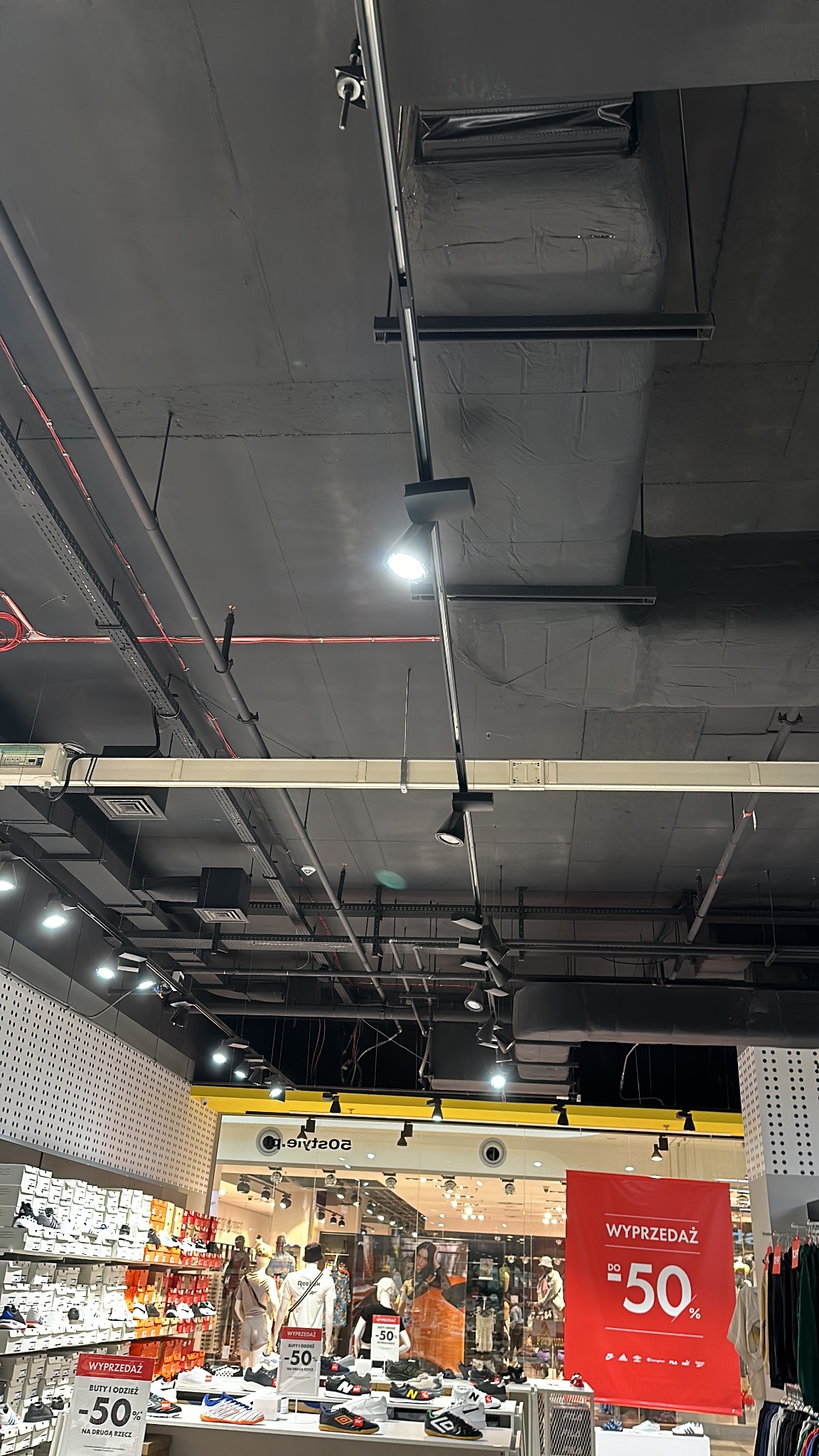 Widok na sufit sklepu z widocznymi instalacjami oświetleniowymi, przewodami i elementami wentylacji, z perspektywy ekspozycji obuwia i odzieży przecenionej.